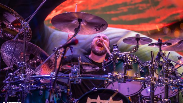 Alex Bent, drummer of Trivium, performing live on stage with full drum kit and energetic stage lighting.