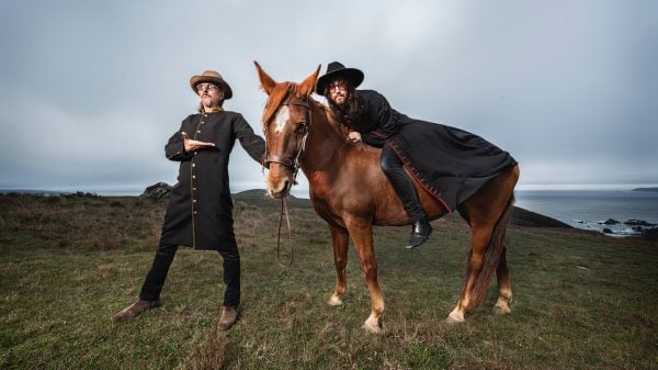 Les Claypool standing next to a horse while Sean Lennon sits on the horse, band The Claypool Lennon Delirium posing outdoors