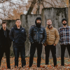 Members of August Burns Red standing outdoors in a fall setting, posing together in front of weathered concrete walls with fallen leaves on the ground.