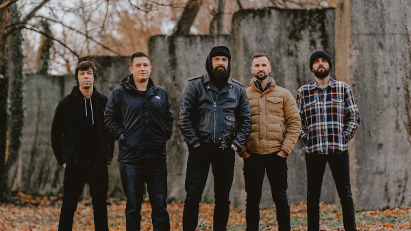 Members of August Burns Red standing outdoors in a fall setting, posing together in front of weathered concrete walls with fallen leaves on the ground.