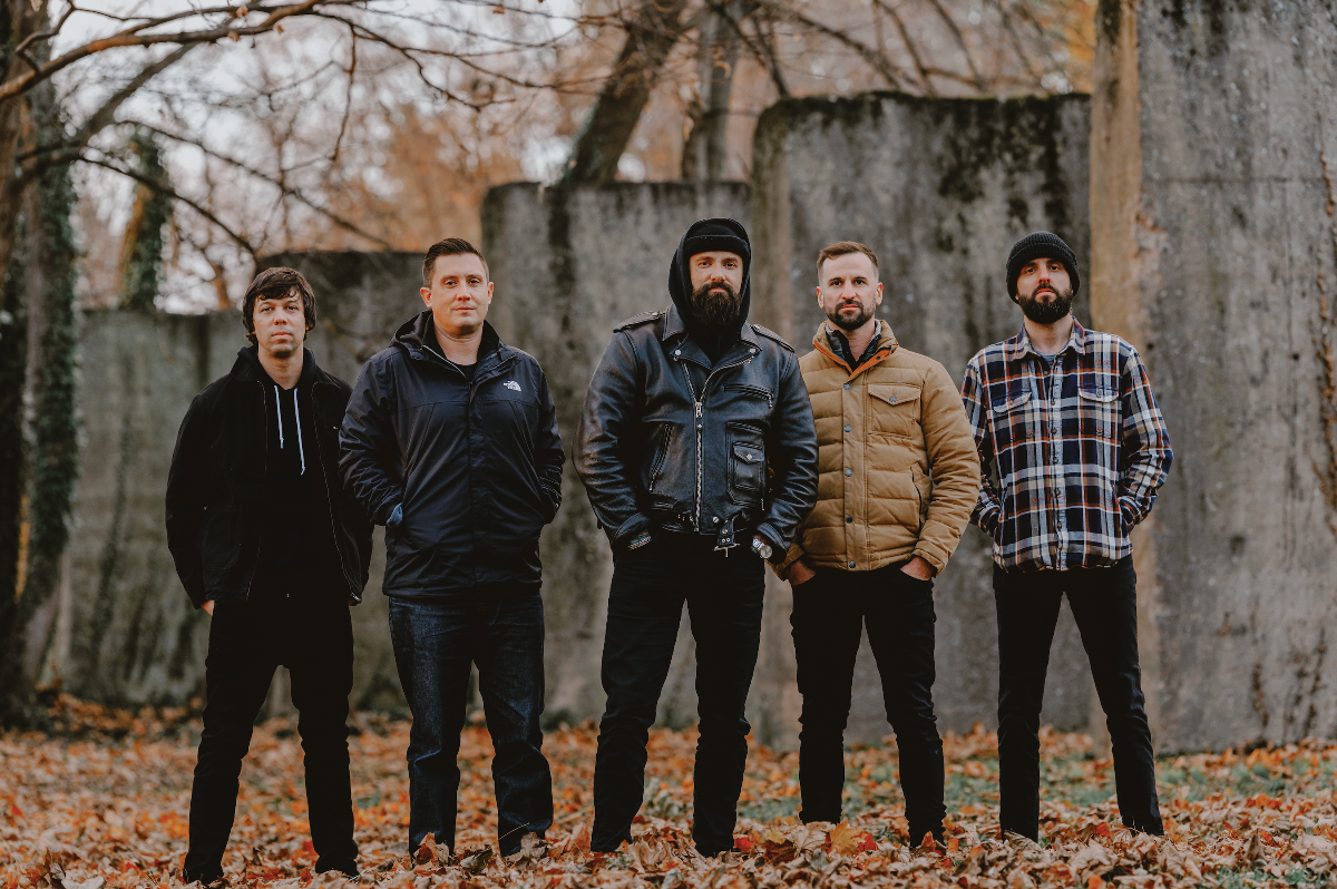 Members of August Burns Red standing outdoors in a fall setting, posing together in front of weathered concrete walls with fallen leaves on the ground.
