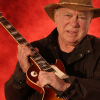 William Shatner holding a Gibson Les Paul guitar, wearing a hat, posed against a bold red background.
